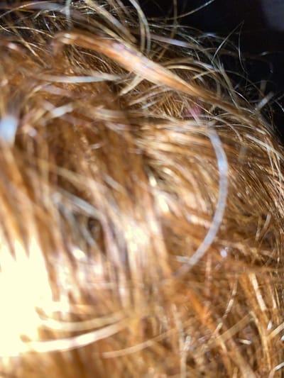 The image is a very close-up, macro shot of human hair. The hair appears to be a light brown or reddish-brown color, with some strands catching the light and appearing lighter or almost white. There are also some very thin, almost transparent strands visible, which could be flyaways or very fine hairs. The focus is very shallow, with parts of the hair being sharp and in focus, while other areas are blurred and out of focus. The lighting creates highlights and shadows within the hair, giving it a dynamic texture. The background is dark and indistinct, suggesting it might be out of focus or a dark surface. There are no people, objects, or environmental details visible that would indicate a specific location within the city of Wumat, Nigeria, or any other location. The image solely focuses on the texture and color of the hair itself.