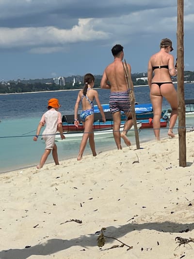 A sandy beach scene in Zanzibar City, Tanzania, shows four individuals walking barefoot from left to right along the shoreline under a cloudy sky. The foreground consists of light-colored sand with scattered dark debris. From left to right: a young child in an orange bucket hat and a light-colored rashguard; a young girl with dark hair in a ponytail wearing a blue patterned one-piece swimsuit; a bare-chested man in blue and white striped swim trunks carrying a long wooden pole over his right shoulder; and a woman in a black bikini top and thong bottom holding a brown bottle in her right hand. A blue rope stretches horizontally across the mid-ground, and two thick, light-colored wooden poles stand vertically on the right. Behind the individuals, several small, brightly colored boats (red, blue, white) are partially visible in clear, shallow turquoise water. The water extends into a deeper blue, with a distant coastline visible across the bay, featuring green vegetation and a city skyline with several multi-story buildings.