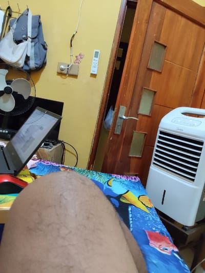 A person's bare leg, from the knee down, is visible in the foreground of the image. The leg rests on a colorful bedspread with blue, red, and yellow designs that resemble an underwater scene. The bedspread lies on a bed that is in front of a white portable air conditioner unit. The air conditioner is located next to a closed, wooden door. The door has a metal handle with a small decorative knob. Behind the air conditioner and to the left is a black laptop computer, with an open screen displaying a white background with text and graphics. The laptop is on a small, black surface with a white-and-gray fan to the left. There is also a gray backpack hanging on the wall behind the laptop. The wall is yellow. There is a small white remote control on the wall. The scene is located in Sidoarjo, Indonesia (latitude: -7.448, longitude: 112.714).