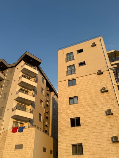 This is a media file featuring two adjoining apartment buildings viewed from a low angle against a clear blue sky. The buildings appear to be in a sunny, warm climate. The time of day is likely late afternoon, judging by the warm, golden light illuminating the facades and casting long shadows.
The building on the left, in the foreground and closer to the viewer, is modern with a grey and beige facade. It features multiple balconies with black metal railings and angular white overhangs. Several windows are visible. Two flags, one red and one blue, are hung on a balcony.
The building on the right, further away, has a cream-colored facade with a textured finish, resembling light-colored bricks. It has numerous windows, some with metal grates. Several outdoor air conditioning units are mounted on the exterior walls of both buildings. The buildings are tall, suggesting they are apartment complexes or hotels in a coastal or urban setting.
There are no people visible in the frame. The scene is calm and peaceful, with the focus on the architecture and the interplay of light and shadow. The location context provided is Rafailovići, Montenegro. No visible text is present in the image.