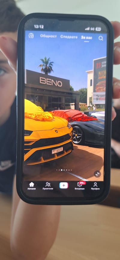 The image shows a person holding a smartphone displaying a TikTok video or photo. The screen is tilted to the side, but the visible content includes a yellow Lamborghini and a dark-colored Ferrari, or a similar exotic car, parked outside a building with the large, illuminated sign "BENO" on it. A partial banner for "Stay Joe" is visible in the background. The license plate of the Lamborghini shows the number "52270." The phone's interface has TikTok icons such as "Начало" (Home), "Приятели" (Friends), "+" (Add), "Входящи" (Inbox), and "Профил" (Profile), all in Bulgarian. The phone's display also shows "Общност" (Community), "Следвате" (Following), and "За вас" (For You). The 5G connection and time, 12:12, are visible at the top left. The photo is captured in Pernik, Bulgaria, where the BENO building is located.
