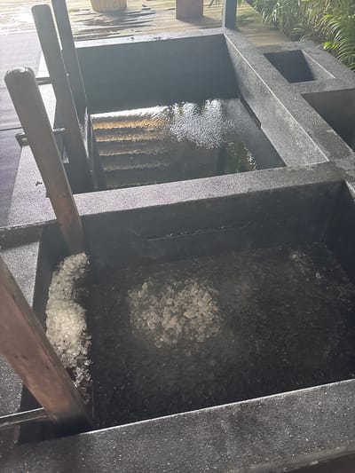The image shows a close-up view of two large, dark, rectangular basins. The basin in the foreground is filled with a layer of crushed ice. A wooden paddle is partially submerged in the ice, suggesting recent use. The basin in the background contains dark water with ripples, indicating movement. The basins appear to be made of a speckled, rough-textured material. The scene is outdoors, with a wooden deck visible in the background. Lush green foliage can be seen to the right, suggesting a tropical or semi-tropical environment. The lighting suggests it is daytime. The location is identified as Ban Bang Rak, Thailand.