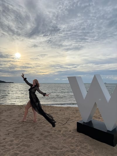 A woman with long, blonde hair is dancing on a sandy beach in front of the ocean. She is wearing a sheer black dress with fur accents on the sleeves. The sun is setting behind her, casting a warm glow on the water. A large white "W" sculpture is positioned to her right. The sky is filled with dramatic clouds. The location is identified as Ko Samui District, Thailand.