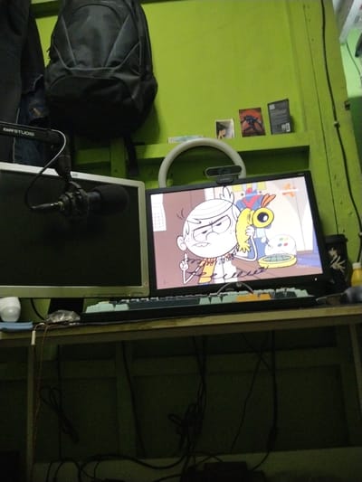 The media file depicts a close-up, slightly low-angle shot of a desk setup with two computer monitors in a room with a lime green wall. On the left monitor, a black microphone with a foam windscreen is mounted on a stand. The left monitor is a silver flat-screen display. To the right of it, a black flat-screen monitor displays an animated character, a young boy with white hair and an orange shirt, holding a yellow object to his ear. He has an angry expression and is pointing his finger. Behind him, a robot and a yellow device with buttons are visible. A ring light is positioned above the monitors, with a webcam attached to it.
To the left of the monitors, a black backpack is hung on a hook on the wall. Above the monitors, on a shelf, are three small items: two framed pictures or cards and a black object with text. The desk surface is cluttered with a keyboard, a white object resembling a mouse or speaker, and various cables.
The scene appears to be indoors, with artificial lighting. The overall mood is somewhat chaotic due to the visible wires and the packed desk space, but the animated content on the screen adds a touch of vibrancy. There is text visible on the microphone stand that reads "taffSTUDIO". The location context indicates Bandung City, Indonesia.
