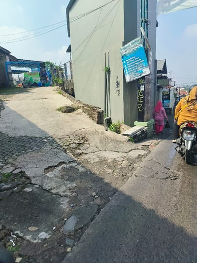 The image shows a narrow, cracked, concrete street in Bandung, Indonesia, located at coordinates -6.907 latitude and 107.704 longitude. The street curves to the right, with a large green building to the right. A blue sign on the building advertises "Toko Sabun," which translates to "Soap Shop" in English. The street is partially shaded by the building's shadow. The street is damaged and cracked, with a small patch of cobblestones visible in the foreground. A woman in a pink hijab is walking down the street towards the left, and a person on a motorcycle is turning left from the street onto a side street. The motorcycle has a license plate with "9256 A" on it. A second person in yellow clothing is behind the motorcycle, walking toward the right side of the image. There is a small, low wall to the right of the street, and a sign that appears to be advertising the soap shop. There are several power lines above the street. The sky is blue and clear, with a few clouds. The scene is typical of a narrow, residential street in an urban area.