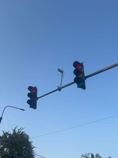 Two traffic lights, both displaying red, are visible against a clear blue sky. The traffic light on the left is positioned lower, with a curved pole extending from the left edge of the frame, likely a street lamp. The traffic light on the right is higher and extends from a horizontal pole. A small surveillance camera is mounted on the horizontal pole between the two traffic lights. Trees are visible at the bottom of the frame, suggesting an outdoor, possibly urban environment. There are no people or animals in the image. The scene is static, with the traffic lights indicating a stop. The overall impression is one of urban infrastructure against a tranquil sky.