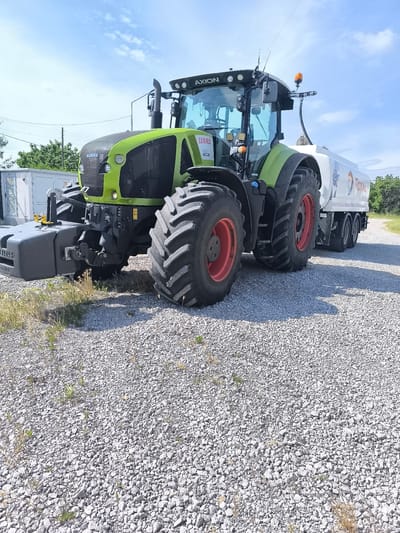 The image displays a bright, sunny day in Inovo, Bulgaria, with a large green and black CLAAS AXION tractor parked on a gravel surface. The tractor has a large white tanker trailer attached to its rear, which is branded with the "TOTAL" logo and other text. The tractor's tires are large and have red rims. In the background, there are some trees and a cloudy blue sky. To the left of the tractor, there is a white electrical box. The scene appears to be a rural or industrial area.