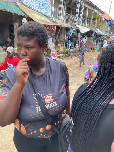 This image depicts a bustling outdoor market scene in Umuode, Nigeria, under bright daylight. In the foreground, a woman with short, dark curly hair looks down, holding a small, light-colored item in her hand. She wears a dark gray t-shirt featuring "999" and "VINES" text, paired with dark leggings adorned with orange and yellow patterns. A black shoulder bag is slung across her body. To her right, another individual with long, dark braided hair is partially visible from behind, wearing a dark top and colorful patterned shorts.
The background showcases a vibrant marketplace filled with numerous people, market stalls, and goods. Various products are displayed, including bottled drinks, fresh produce like onions and tubers, and fruits. Multi-story buildings, some featuring distinctive black and white checkered patterns on their lower facades, line the street. Several signs are visible, prominently displaying "POS" in red lettering, along with "TONY BEST." Another sign includes contact details: "+2347064668924", "+2349038803472", and mentions "BUSINESS ID BROCHURES ITC" and "ARIARIA INTERNATIONAL MARKET ABA, ABIA STATE". The scene conveys an active and energetic commercial environment.