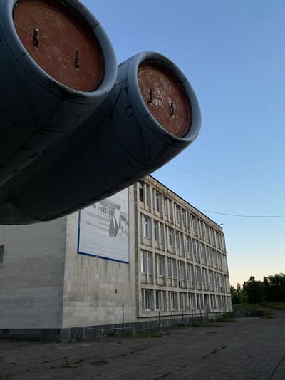 The image shows a low-angle view of two large, dark cylindrical objects in the foreground, appearing to be jet engine nacelles. Their interiors are a reddish-brown color, and they show signs of wear and tear. Behind these objects, a tall, concrete building with numerous windows stretches upwards. A large banner with a black and white image and text in Russian is affixed to the building's facade. The banner depicts what appears to be a group of people in formal attire. A paved walkway runs alongside the building on the right side of the image. The sky above is clear and a light blue, suggesting daytime. Trees are visible in the distance behind the building. The scene is likely an outdoor setting in Ulyanovsk, Russia, possibly near an aviation-related museum or monument, given the prominent display of what look like aircraft engines. The overall impression is one of weathered industrial elements and a somber architectural style.