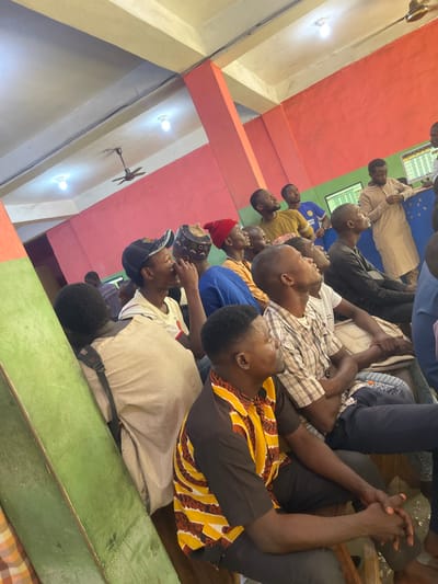 The image displays an indoor scene in Zaria, Nigeria, featuring a group of approximately twenty men, predominantly young adults. They are seated on the floor, arranged in rows, with most individuals looking upwards and towards a focal point outside the frame, suggesting attentive observation or listening. Many have their heads tilted back, and some have their hands clasped, indicating engagement.
The men wear a mix of clothing styles, from casual t-shirts and plaid shirts to more traditional attire. One man in the foreground wears a distinct brown and yellow patterned long-sleeve shirt. Another sports a blue t-shirt with "ENI" partially visible, and a cap with "NE" visible is worn by a man in the bottom right.
The interior space features walls painted in contrasting colors of red and green, alongside beige pillars. Overhead, a ceiling fan and two light fixtures provide illumination. In the background, two wall-mounted screens display text or data that is unreadable from the distance, hinting at a setting used for presentations, education, or monitoring information. The general atmosphere is one of calm attention and communal gathering, possibly for a lecture, discussion, or religious event. The bright lighting suggests it is either daytime indoors or a well-lit evening event.