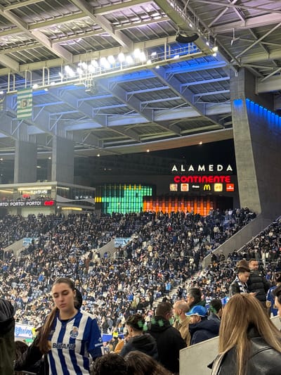 An angled view of a packed stadium at night. Bright lights illuminate the metal framework of the roof on the left. A large scoreboard displays advertisements for Alameda, Continente, McDonald's, and others. A green and yellow pixelated design shines below the advertisements. A sea of spectators fills the stands, with a person in a blue and white striped jersey prominent in the foreground. Coca-Cola advertisements can be seen near the bottom of the image.