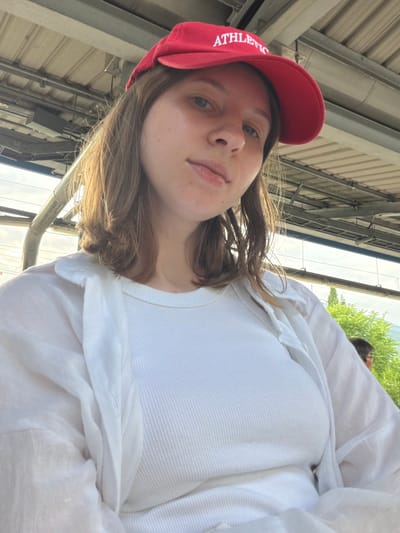 A young woman wearing a red baseball cap with "ATHLETIC" embroidered in white sits outdoors, appearing to be at a train or subway station in Seoul, South Korea. She is wearing a white ribbed tank top and an unbuttoned white shirt over it. The background shows the station's overhead structure and some greenery. The image is a close-up, focusing on the woman's face and upper body.