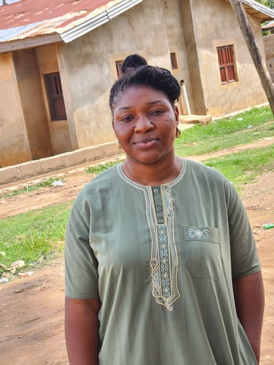 The image depicts a person, likely of African descent, standing outside in what appears to be a rural or semi-rural setting. The person is wearing a long-sleeved, light green garment with intricate embroidered patterns around the neckline and down the front. The garment has a pocket on the right side, also adorned with embroidery. The person's hair is dark and styled in a bun.
The background reveals a simple building with light-colored walls and small, dark-framed windows. Adjacent to the building, there is an area covered with green grass and some patches of dirt. The ground in the foreground is also dirt. There's a rusted corrugated metal roof visible on the left side of the image, suggesting it might be part of another structure or an extension.
The scene appears to be outdoors during the daytime, with natural lighting. The overall impression is casual and everyday. No specific activity or event is clearly taking place; it seems to be a posed or candid portrait. The person's expression is neutral, and their gaze is directed towards the camera.
Location cues like the building style, vegetation, and the general landscape suggest a location in Sabon Tasha, Nigeria, as provided. The weather appears to be fair. There is no visible text in the image.