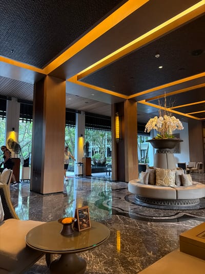 The image depicts the spacious and elegantly designed lobby of a hotel in Kuta, Indonesia. The lobby features a high ceiling with intricate dark patterned panels and illuminated with bright orange linear lights, creating a modern and luxurious ambiance. Large wooden pillars support the ceiling, and the floor is covered with polished dark marble, reflecting the surrounding light and decor.
In the center of the lobby, a circular, plush seating arrangement is placed around a large decorative planter filled with white orchids. Several decorative pillows with ornate patterns are scattered on the seating. To the left, a small round coffee table holds a decorative bowl with orange flowers and a framed menu or informational sign.
In the background, large glass windows and doors offer views of lush greenery and outdoor trees, suggesting an open and tropical setting. Two individuals are visible in the lobby. One person, wearing a white t-shirt and shorts, is walking towards the right side of the frame. Another person, wearing a black top and shorts, is walking towards the left.
The overall impression is that of a high-end hotel lobby, likely designed to provide a welcoming and sophisticated experience for guests. The combination of natural elements, modern design, and subtle lighting creates a serene and inviting atmosphere.