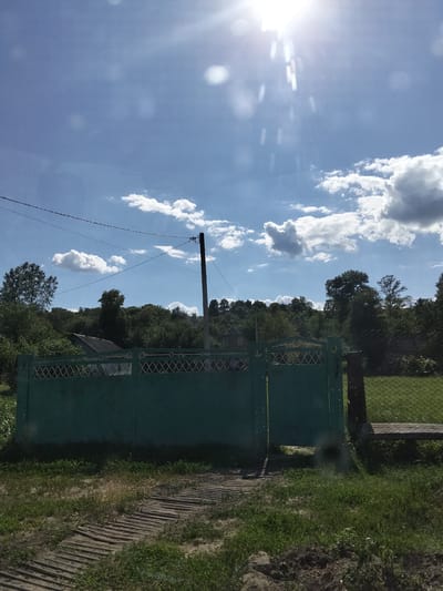 The image depicts a sunny day in Ustyvytsia, Ukraine, with a clear blue sky filled with scattered white clouds. The sun is bright and positioned high in the sky, creating lens flare. In the foreground, a green metal gate stands open, with a decorative lattice pattern along its top. To the left of the gate, a wooden walkway is laid out on the ground. Behind the gate, lush green trees and foliage form a dense backdrop, with a hint of a house visible amongst the trees. A power line stretches across the sky, supported by a utility pole on the right side of the frame. A chain-link fence runs along the right edge of the image, bordering a grassy area. The overall scene suggests a rural or semi-rural setting on a pleasant day.