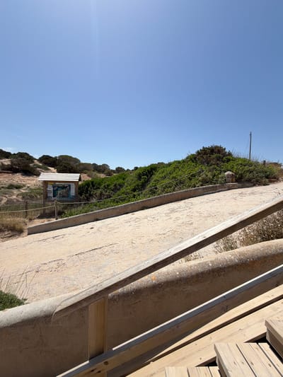 A bright, sunny daytime, low-angle shot captures a pathway leading up a gentle incline. The pathway is made of a sandy, concrete material and is bordered by a weathered concrete wall and wooden railings. To the left of the pathway, a short wooden fence encloses a patch of dry grass and scrub. Beyond the fence, a wooden information board stands under a sloped roof, displaying maps and text, suggesting a park or natural reserve. The background is filled with dense, green vegetation and a clear blue sky. The scene is located in the municipality of Sant Josep de sa Talaia, Spain.