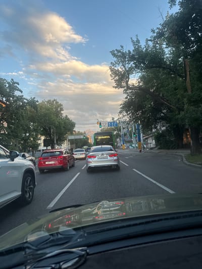 The image depicts a traffic scene in Almaty, Kazakhstan, on a cloudy day. The perspective is from inside a vehicle, looking forward through the windshield. Several cars are visible on the multi-lane road, including a white SUV in the foreground on the left, a red Hyundai in the middle, and a silver Kia on the right. A green bus with "19" displayed on its front is also in traffic. Traffic lights are visible, with some showing green. Tall trees line the sides of the road, and buildings can be seen in the background. The sky is partly cloudy, with patches of blue and illuminated clouds. The overall atmosphere suggests a typical urban commute during daytime.