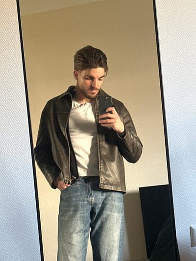 An image captured in Bertrange, Luxembourg, depicts a male individual taking a mirror selfie indoors. The subject, a male with styled light brown hair and facial stubble, is reflected from the mid-chest down in a large, rectangular mirror. He wears a dark brown leather jacket over a light-colored Henley-style t-shirt, light blue denim jeans, and a dark belt. His right hand holds a dark-colored smartphone, positioned to capture the reflection, while his left hand is placed in his left jeans pocket. His gaze is directed downwards towards the smartphone/reflection. The background reflected behind the subject consists of a plain, light beige wall. To the immediate left of the mirror, a textured, off-white wall is visible. To the right, another plain, light-colored wall is seen, with a partial reflection of a dark, rectangular object in the lower right corner. The lighting source appears to be artificial, casting a warm hue over the scene.