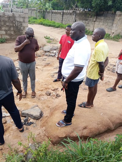 In Zango-ndaji, Nigeria, a group of five men stands on a sandy, uneven terrain that appears to be a road or pathway. The ground is littered with debris, including rocks and what looks like broken asphalt. A low, weathered brick wall runs along the background, with dense green foliage and trees visible beyond it.

The man in the foreground, on the left, is wearing a dark grey t-shirt and dark blue trousers. He is facing away from the camera, with his left hand extended. Next to him, a man in a dark brown t-shirt and grey sweatpants looks down thoughtfully, resting his chin on his hand. He is wearing sunglasses.

To his right, a man in a red t-shirt with "BOSS" printed on it stands with his arms behind his back. He is looking to his left. Beside him, a man in a white polo shirt with blue and black stripes on the collar and sleeves is the most prominent figure. He is looking forward and has his arms clasped behind his back. He is wearing black trousers and blue sandals. Further to the right, a man in a yellow t-shirt and dark patterned shorts stands with his arms behind his back, also looking forward. His grey sandals are visible. Another individual, partially visible in the background on the far right, wears a red t-shirt and light-colored shorts.