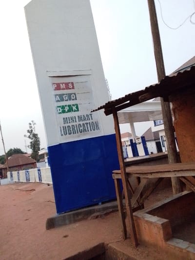 This image depicts a petroleum fueling station located in Ankpa, Nigeria. In the foreground, on the left, a tall, white and blue rectangular sign is visible. The white upper section of the sign features three labeled rectangular panels for fuel types: "PMS" (red letters), "AGO" (green letters), and "DPK" (green letters). Each panel has an empty slot for price display, indicated by a dollar sign next to "PMS." Below these, the words "MINI MART" and "LUBRICATION" are stenciled in black. The lower half of the sign is solid blue.
To the right of the sign, partially obscuring the view, is a rustic, makeshift structure. This structure consists of wooden posts supporting a corrugated metal roof and a rough wooden shelf. Below this shelf, a small, unrendered brick or concrete structure with a square opening is present at ground level.
The ground in the immediate foreground and extending to the left appears to be red-brown earth or dirt, with some unevenness and shallow depressions. Behind the main sign, a low white fence with blue accents runs horizontally, separating the station area from what appears to be a dusty road. In the background, beyond the fence and a tree, several single-story buildings with reddish roofs are visible, consistent with local architecture. To the far right, part of the main fueling station canopy, supported by white and blue striped pillars, can be seen. A wooden utility pole with wires runs vertically on the right side of the image, near the makeshift structure. The sky is overcast and light. No people or vehicles are visible.