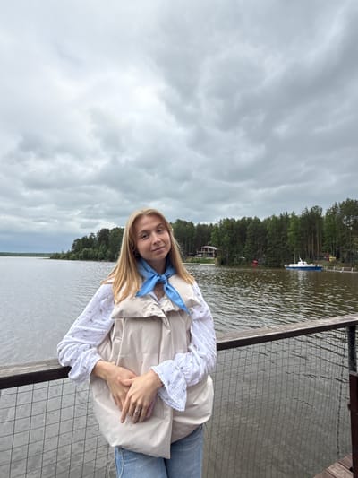 A young woman with blonde hair, wearing a white blouse, a beige puffer vest, and blue jeans, stands on a wooden pier next to a body of water. She has a blue scarf around her neck and is holding a pink object in her hands. The pier has a metal mesh railing. In the background, across the water, is a dense forest with a few buildings visible, including a house and a boat docked. The sky is overcast with grey clouds. The location is the Kama River in the city of Chaykovsky, Russia.