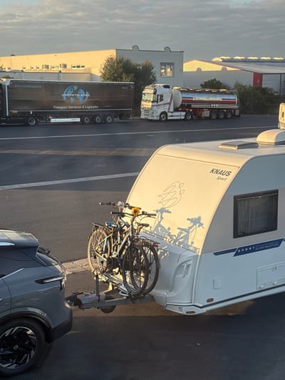A grey crossover SUV is shown from the rear-left, hitched to a white Knaus Sport Silver Selection caravan. Two dark-colored bicycles are mounted on a rack attached to the tow hitch between the vehicles; a yellow sunflower is affixed to the handlebars of the front bicycle. The caravan features "KNAUS Sport" branding, a window on its left side, and a stylized graphic on the upper rear panel, illuminated by low-angle sunlight that casts elongated shadows of the bicycles onto the caravan's rear.
In the mid-ground, a dark semi-trailer truck with "CARPATIA SPED Transport Spedycja & Logistyka" branding is parked or stationary. To its right, a white semi-truck pulls a red, white, and blue tanker trailer. Behind these vehicles, industrial-style buildings with light-colored facades and various rooflines are visible under a partly cloudy sky. The entire scene takes place on a paved surface, likely a parking area or service road, in Himmelkron, Germany.