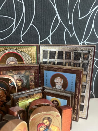The image shows a collection of small, antique religious icons, likely Orthodox Christian, arranged on a white surface. The icons vary in size and design, with some framed in ornate gold and silver detailing, and others appearing to be on wooden bases. Several of the icons depict figures with halos, traditional religious imagery, and some have Cyrillic inscriptions. The background features a dark wall with a repeating abstract pattern of white lines. The overall impression is of a devotional display, possibly a personal collection or part of a larger religious artifact display. There are no people or actions depicted in the image, and the scene is static. The specific location in Pokrovnik, Bulgaria is not discernible from the visual evidence provided.