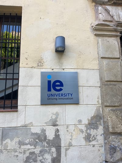 The image shows a close-up view of a building wall in Segovia, Spain, located at latitude 40.953 and longitude -4.119. The wall is made of light-colored stone and is partially covered in peeling paint. A rectangular sign is mounted on the wall, displaying the words "ie UNIVERSITY Driving Innovation" in blue lettering on a gray background. A small, cylindrical light fixture is mounted on the wall above the sign. The sign is positioned on the wall, slightly to the left of the center. A window is located to the left of the sign and is obscured by a metal grate. There are no people or other objects present in the scene. The image is taken in an outdoor setting, as evidenced by the presence of the wall and the light fixture.