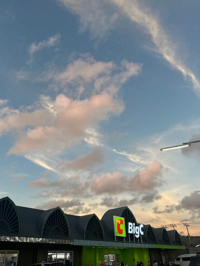This media file captures a low-angle view of the sky and the exterior of a "Big C" supermarket in Ko Pha Ngan, Thailand. The sky is a prominent feature, filled with a mix of light blue and scattered clouds, some of which are tinged with pink and orange, suggesting either sunrise or sunset. Wispy clouds streak across the upper portion of the image, while more voluminous, textured clouds are present lower down. A long, fluorescent light fixture is visible against the sky in the upper left quadrant, attached to a pole.
The supermarket building occupies the right side of the frame. It has a modern architectural design with a dark, curved roof structure that is segmented by fan-like awnings over windows. The prominent "Big C" logo is displayed in large, illuminated white letters against a dark background, with a smaller, green illuminated sign featuring the "Big C" logo to its left. The building's facade is predominantly green and dark gray. Visible through windows are displays and what appear to be aisles within the store. A white van is parked to the far right, and there are hints of people and motorcycles near the entrance.
The overall impression is a snapshot of a commercial establishment in a tropical setting, with the natural beauty of the sky providing a dramatic backdrop. The time of day appears to be either early morning or late afternoon/early evening, given the colors in the sky. The weather seems fair with scattered clouds. The visible text includes the "Big C" logo.