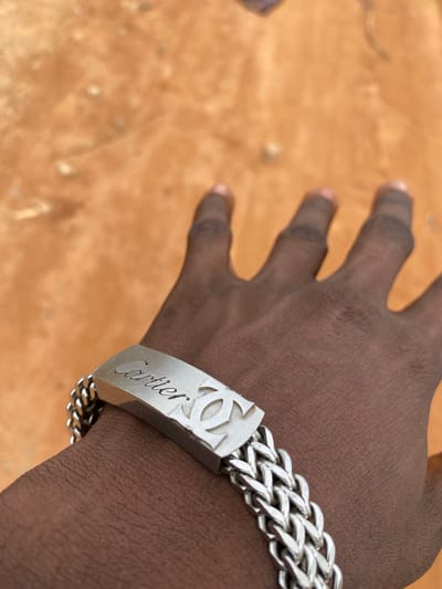 A close-up, medium shot captures a dark-skinned hand with a silver bracelet on its wrist. The bracelet features a rectangular plate engraved with the word "Cartier" in cursive, accompanied by a logo. The hand is positioned on a textured, orange-brown surface that appears to be dirt or sand, possibly a street or outdoor area in Kuson, Nigeria. The background is blurred, with hints of pebbles or small stones scattered across the ground. The lighting suggests an outdoor setting, likely during the daytime. The image focuses on the bracelet and the hand, with the environment serving as a backdrop.