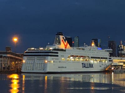 This is a nighttime, long-exposure photograph of a large ferry docked in the city of Tallinn, Estonia. The ferry, named "Tallink," is white with black accents and features multiple decks with illuminated windows. A prominent red and white V-shaped logo is visible on the superstructure of the ship. The ferry is reflecting in the dark water of the harbor, creating bright yellow and orange reflections. On the left side of the image, a pier structure with illuminated lamps is visible. In the background, the illuminated skyline of Tallinn, with several modern buildings, is faintly discernible against the dark blue night sky. A single streetlamp, emitting a warm yellow glow, is positioned to the left of the ferry. There are no people visible in the frame.