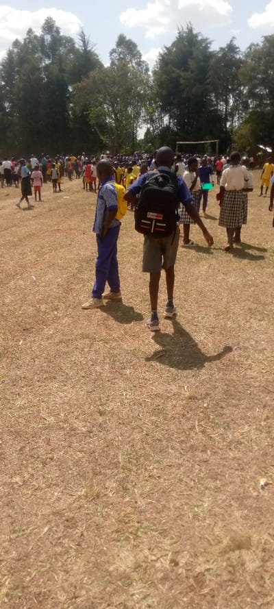 The image shows a large gathering of people in Malava, Kenya, at latitude 0.53 and longitude 34.811. There are many people standing in the background, mostly children, wearing school uniforms. In the foreground, two boys, wearing school uniforms, are walking towards the camera. One boy is wearing a blue shirt and blue pants, and the other is wearing a blue shirt and gray shorts, and carrying a black backpack with the word "Fashion" written on it. The ground is dry and covered with grass and twigs. The sky is partly cloudy.
