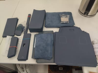 The image shows a collection of dark blue leather or faux leather cutouts, likely for crafting bags or wallets. The cutouts are neatly stacked in various piles, some held together with rubber bands. There are also some pieces of blue suede fabric and a small plastic bag containing what appear to be metal findings or hardware, with text labels. In the background, a stainless steel kettle is visible, and a white table surface serves as the setting. The scene appears to be a workspace, possibly a home studio or a small workshop in Hrodna, Belarus. The lighting suggests it could be daytime. The overall impression is one of preparation for a crafting project. Visible text on the labels includes "MAKEУ", "BAGS", and some Cyrillic characters that are partially obscured or difficult to read, but appear to be related to product descriptions or manufacturing details. Another label with Cyrillic text is visible on a document next to some of the cutouts.