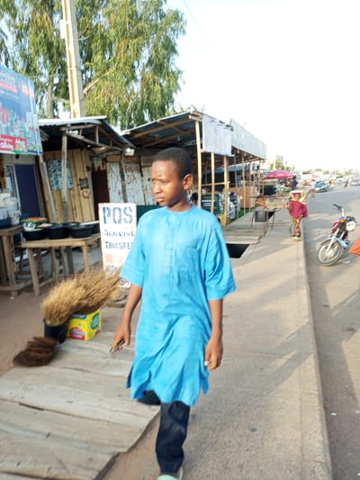 In Tempure, Nigeria, a young man in a bright blue traditional tunic walks along a sun-drenched street lined with market stalls. He walks from left to right in the frame, his gaze directed forward. Behind him, a variety of wares are displayed on wooden tables. One stall features black bowls filled with food, and a prominent "POS SERVICE TRANSFER" sign is visible. To the left of the man, a broom made of dried grass stands upright in a yellow box. Further down the street, a woman in a maroon headscarf and patterned trousers is seen near a red umbrella. A red motorcycle is parked on the right side of the road, its silver spokes catching the light. The environment appears to be a bustling marketplace under a bright, clear sky.