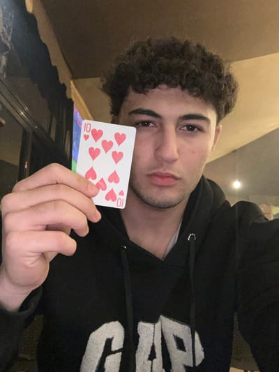 A young man with curly hair holds up a playing card, the ten of hearts. He is wearing a black hoodie with white lettering. The background is slightly blurred, suggesting an indoor setting with neutral-toned walls. The man looks directly at the camera with a serious expression. The image is taken at a slight angle, lending a casual, selfie-style feel.