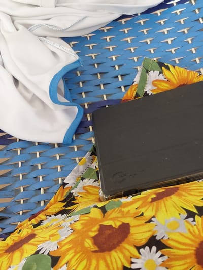 A close-up shot captures a black object, possibly a book or tablet, lying on a blue woven surface. To the left of the black object, a white garment with a blue trim is draped, showing a textured fabric. In the foreground and partially obscured by the black object and white garment, a colorful fabric with a sunflower print is visible. The blue woven surface appears to be part of an outdoor chair or lounge. The scene is brightly lit, suggesting an outdoor setting. There is no discernible information about the city of Santa Ana, Venezuela, in this image.