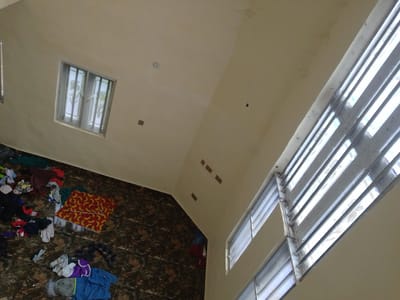 The image depicts an interior scene, likely within a residential building in Obosi, Nigeria. The perspective is from a high angle, looking down onto a tiled floor and up towards a sloped ceiling.
On the floor, scattered clothing items are visible. These include a red and yellow patterned cloth, various t-shirts, shorts, and other apparel in different colors such as blue, purple, grey, and black. Some items appear to be folded, while others are in disarray. The floor itself is covered in dark, glossy tiles with a marbled pattern.
The walls are a light, neutral color. A window with vertical bars and reflective glass is situated on one wall, allowing light to enter the room. Another window, also with horizontal blinds, is visible on a higher plane, suggesting multiple levels or an open-plan design. The ceiling is a pale, cream color and shows some signs of minor imperfections or water stains in places. Several square ventilation grates or electrical boxes are embedded in the ceiling. In the upper part of the image, a circular object, possibly a smoke detector or light fixture, is mounted on the ceiling. The overall impression is of a somewhat untidy but functional living space.