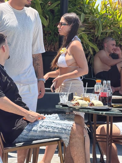 The image captures a casual outdoor gathering, likely during the day, given the bright sunlight and shadows. In the foreground, a person wearing a white t-shirt is partially visible, lying down. To their right, another person in a black t-shirt is also lying down. In the middle ground, a woman in a white bikini and a crocheted cover-up is seated, holding a phone. She is wearing sunglasses and has a flower accessory in her hair. Behind her, a man is lying down with his torso exposed, wearing black shorts. A table with drinks, including several glass bottles of Coca-Cola and a bottle of water, is visible. There are also some food items on the table, possibly wraps or burritos. The setting appears to be a relaxed outdoor environment with lush green foliage and a hint of water in the background, suggesting a poolside or garden area. The overall mood seems laid-back and social. There are no discernible text elements in the image.