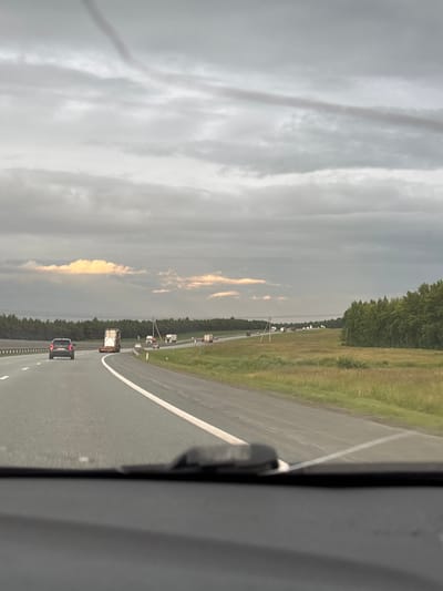 The image captures a view from inside a vehicle, looking out at a highway scene in Maksimov Pochinok, Russia. The orientation is vertical, suggesting the photo was taken with the camera held sideways. The foreground is dominated by the interior of the vehicle, with a dark grey pillar on the right side. The highway itself is visible, with a white lane marker clearly defining the road. Several vehicles, including cars and trucks, are traveling in the same direction on the highway.

The roadside is lined with a grassy verge and a dense forest in the distance. The sky is overcast with dramatic clouds, some of which are illuminated by what appears to be sunset or sunrise light, casting a warm glow. The weather appears to be cloudy, possibly indicating an impending rain shower or a recent one, as the road surface seems slightly damp in places. The overall mood is one of travel and transit, with the vastness of the sky and the open road suggesting a journey. There is no visible text in the image.
