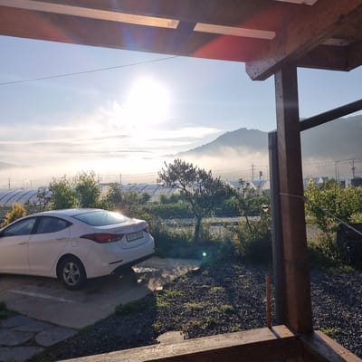 This image depicts a tranquil morning scene in Dunnae-myeon, South Korea. A white sedan is parked in a gravel and concrete area, with its rear facing the camera. The car's license plate reads "41라 5250." In the background, rolling hills are shrouded in a soft mist, with the sun rising brightly, casting a warm glow. Several greenhouse structures with arched roofs are visible, suggesting agricultural activity. A sparse tree with dark foliage stands near the car, and other vegetation, including bushes and grasses, are scattered throughout the foreground and mid-ground. The scene is framed by a wooden structure, possibly a porch or a building overhang, in the foreground. The overall atmosphere is serene and rural, indicative of early morning in a countryside setting.