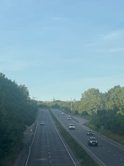 An aerial view captures a multi-lane highway slicing through a verdant landscape under a clear, pale blue sky. The highway, paved with asphalt, features white dashed lines demarcating the lanes and solid white lines along the edges. Vehicles, including cars and a white van, are visible on the road, traveling in both directions. Lush green trees line both sides of the highway, their dense foliage creating a natural border. In the distance, beyond the trees, industrial structures including tall power line pylons and a chimney or smokestack are visible against the horizon. The scene appears tranquil, with the sunlight casting a soft glow on the environment. The location is identified as Borough of Swale, United Kingdom.