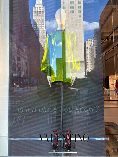 This is a photograph of a store window display in New York City, United States of America, likely on Fifth Avenue or a similar upscale shopping district. The display features a mannequin dressed in a bright, translucent yellow and blue garment adorned with embellishments around the neckline. The mannequin is positioned in front of a digital screen that projects an image of the New York City skyline, complete with tall buildings and a bright blue sky with clouds. Below the mannequin, text that reads "Is it a place you know?" is displayed on the screen. At the bottom of the display, a pair of red, studded high-heeled shoes are showcased next to the "VALENTINO" logo. The overall scene conveys a sense of luxury and urban sophistication, typical of high-end fashion retail in New York City.