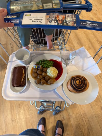 The image depicts a tray of food in an IKEA shopping cart. The tray contains a plate of Swedish meatballs with mashed potatoes, lingonberry jam, and peas, adorned with a small Swedish flag. Additionally, there is a cinnamon roll, a glass of water, a blank white mug, and a dessert resembling tiramisu. A child’s feet are visible in the shopping cart. A woman’s shoes and jeans are visible on the right of the image. The IKEA cart features the company’s logo and website address. There is German text advertising the availability of children’s meals. The image was likely taken at an IKEA in Kaiserslautern, Germany.