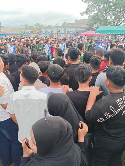 A high-angle, full-shot image captures a dense crowd of people in what appears to be an outdoor event or gathering in Desa Meunasah Blang, Indonesia. The sky is overcast. The foreground is dominated by the backs of several individuals, including a woman in a black hijab looking at her phone, and a man in a white collared shirt. Behind them, a large number of people are visible, many wearing casual clothing. Several individuals in military-style camouflage uniforms are interspersed within the crowd. Structures, including tents and buildings, are visible in the background, suggesting a community or public space. The ground appears to be dirt or grass. The overall impression is a busy, crowded scene with a sense of collective focus, though the specific nature of the event is not clear from the image alone.