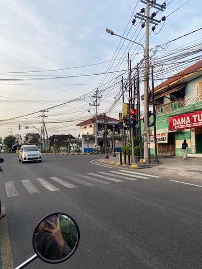 The image depicts a street scene in Madiun, Indonesia, taken from the perspective of a motorcyclist stopped at a crosswalk. A white car with license plate AE 1419 BM is visible ahead. Above, utility poles covered in a tangle of wires stretch along the street. Buildings line the street, including one with signage for "DANA TU," "JAMINAN BPKB M," "SUPERIA," and "TTO KILAT PIGURA." A traffic light is showing red. The side mirror of the motorcycle reflects the motorcyclist's hand holding a cell phone.