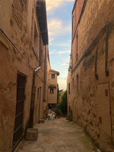 The image shows a narrow, cobblestone alleyway in the historic city of Segovia, Spain, located at latitude 40.952 and longitude -4.129. The alleyway is lined with two tall, ochre-colored buildings, both with exposed stone walls. The building on the left has a large, barred window, while the building on the right has several smaller windows and a closed wooden door. The buildings are weathered and show signs of age. The alleyway is empty of people and there is a pile of debris in the middle of the frame. The sky above the alleyway is blue with a few white clouds. The image was likely taken on a sunny day, as the shadows cast by the buildings are quite short. The alleyway is a typical example of the narrow, winding streets found in many of Spain's older cities. There is an empty cardboard box in the bottom left corner with the word "Tel" written on it. A small bush is growing in the far right corner. A metal downspout runs down the left side of the image, from the roof of the building to the ground.