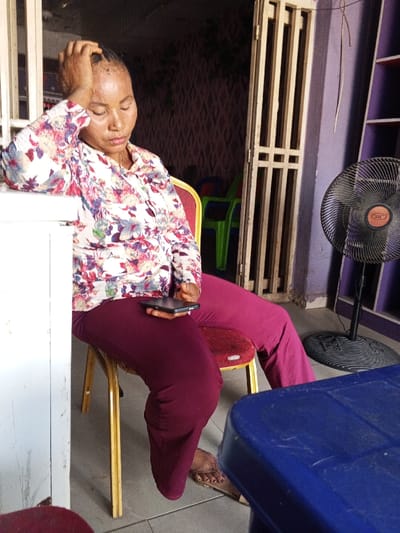 One adult female is seated indoors in Zango-ndaji, Nigeria. She wears a long-sleeved, collared shirt featuring a multicolored floral pattern and solid maroon trousers. Her right elbow rests on a white, rectangular structure, possibly a counter or table, with her right hand positioned on her forehead and eyes appearing closed or downcast. A black smartphone is horizontally placed on her left thigh. She occupies a red-seated chair with gold-colored metal legs. Her feet, clad in brown sandals, are visible on the light-colored floor. In the background, a doorway with light-colored, vertically slatted security bars is visible. To the right of the doorway, a black standing electric fan with an orange circular logo is present. The background also shows patterned wall coverings and other colored plastic chairs in dimly lit areas. A blue plastic table corner is visible in the lower right foreground.