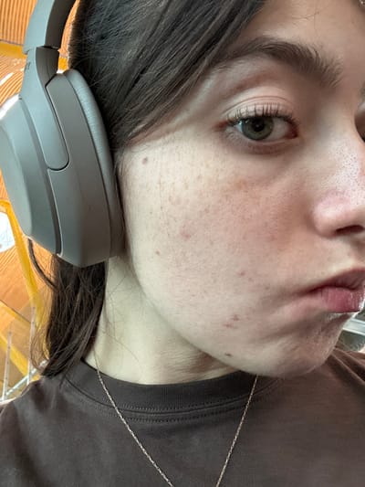 The image presents a close-up portrait of a young individual, appearing female, with dark brown hair partially obscuring the forehead and left ear. She wears light gray Sony over-ear headphones on her left ear. The subject's visible right eye is green, and her light complexion exhibits numerous freckles and several minor epidermal blemishes on the cheek and jawline. Her lips are slightly parted or pursed. A thin, silver-colored chain necklace rests against a dark brown crew-neck t-shirt. The blurred background features distinctive wavy, yellow-orange wooden ceiling structures, identifying the location as Terminal 4 (T4) of Adolfo Suárez Madrid–Barajas Airport in Madrid, Spain.
