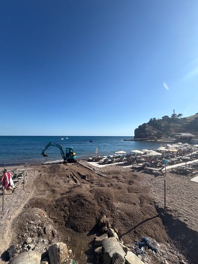 An aerial view captures a sunlit beach scene in Sutomore, Montenegro, under a clear blue sky. The turquoise water of the Adriatic Sea gently laps against the shore. In the foreground, construction work is underway on the sandy beach, with a teal excavator actively moving dark sand. Piles of sand and debris are visible, along with stacked beach chairs and umbrellas in the background, indicating a resort area. A few boats and a jet ski are dotted on the water. A rocky cliffside with some vegetation and structures forms the backdrop to the right of the image, with a building featuring the text "HABANA CLUB" visible near the water's edge. A person in a red and white garment walks away from the camera on the left side of the beach. The overall impression is one of a coastal area undergoing development or maintenance, with recreational elements present.