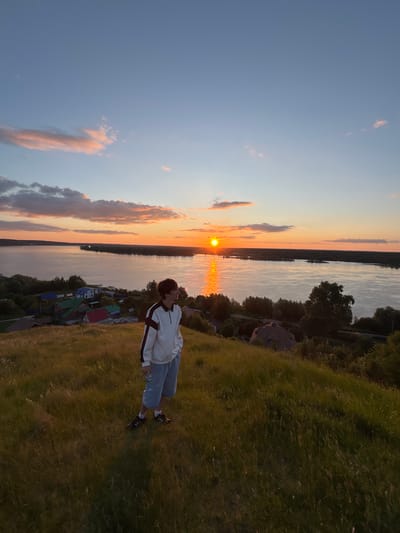 A young person stands on a grassy hill overlooking a wide river during sunset. The person, facing away from the camera and to the right, wears a white jacket with dark stripes on the sleeves and shoulders, light blue shorts, and dark shoes. The sun is setting over the river, casting an orange glow across the water and sky. In the distance, across the river, is a tree-lined bank and a small settlement of houses with colorful roofs. The foreground is dominated by tall, dry grass, suggesting a rural or semi-rural environment. The sky exhibits a gradient of blue and orange, with some wispy clouds catching the sunlight. This scene is located in the village of Olkhovka, Russia.